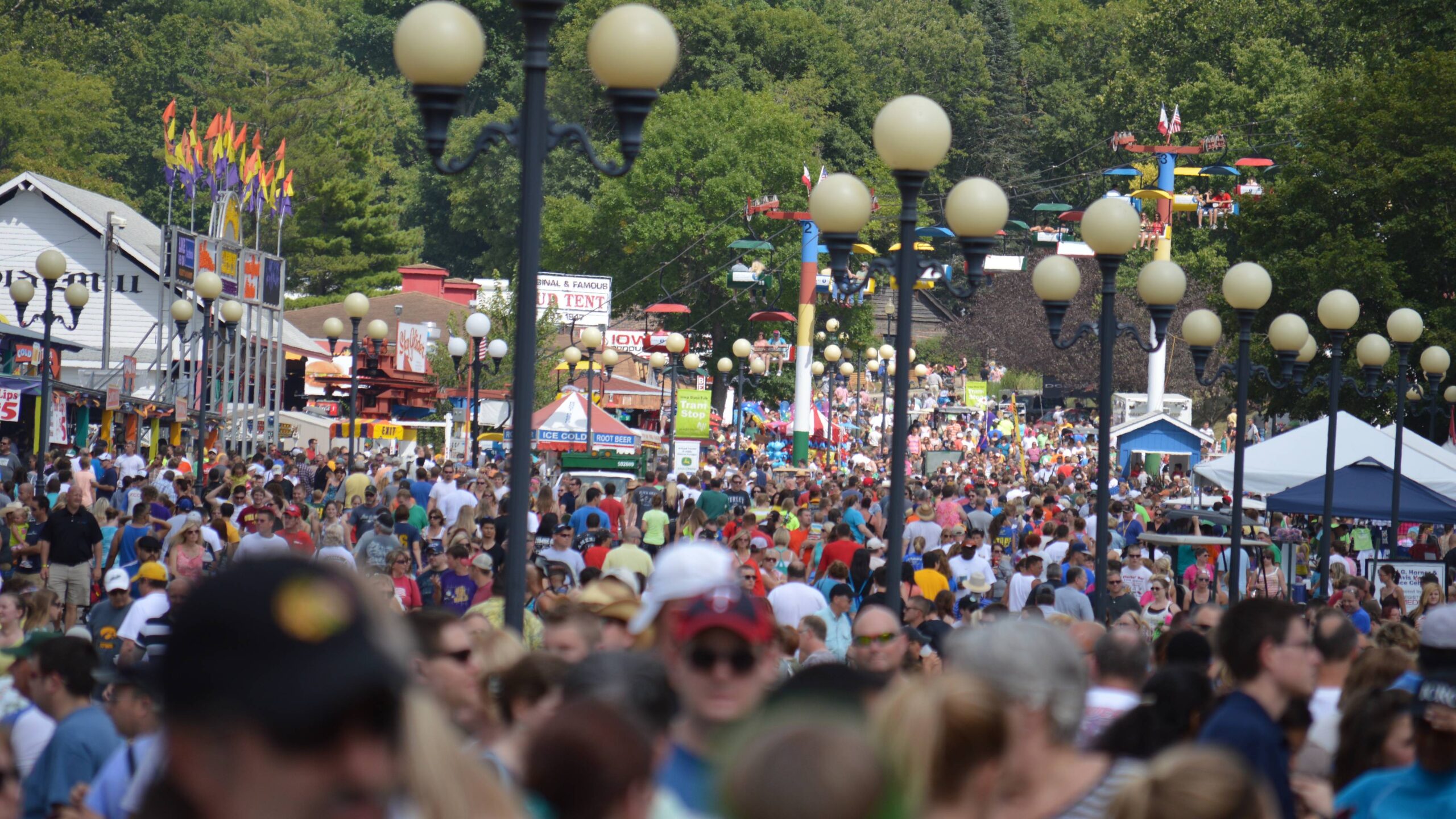 Large crowds at the Iowa State Fairgrounds in Des Moines, with vendor booths and midway rides visible in the background.
